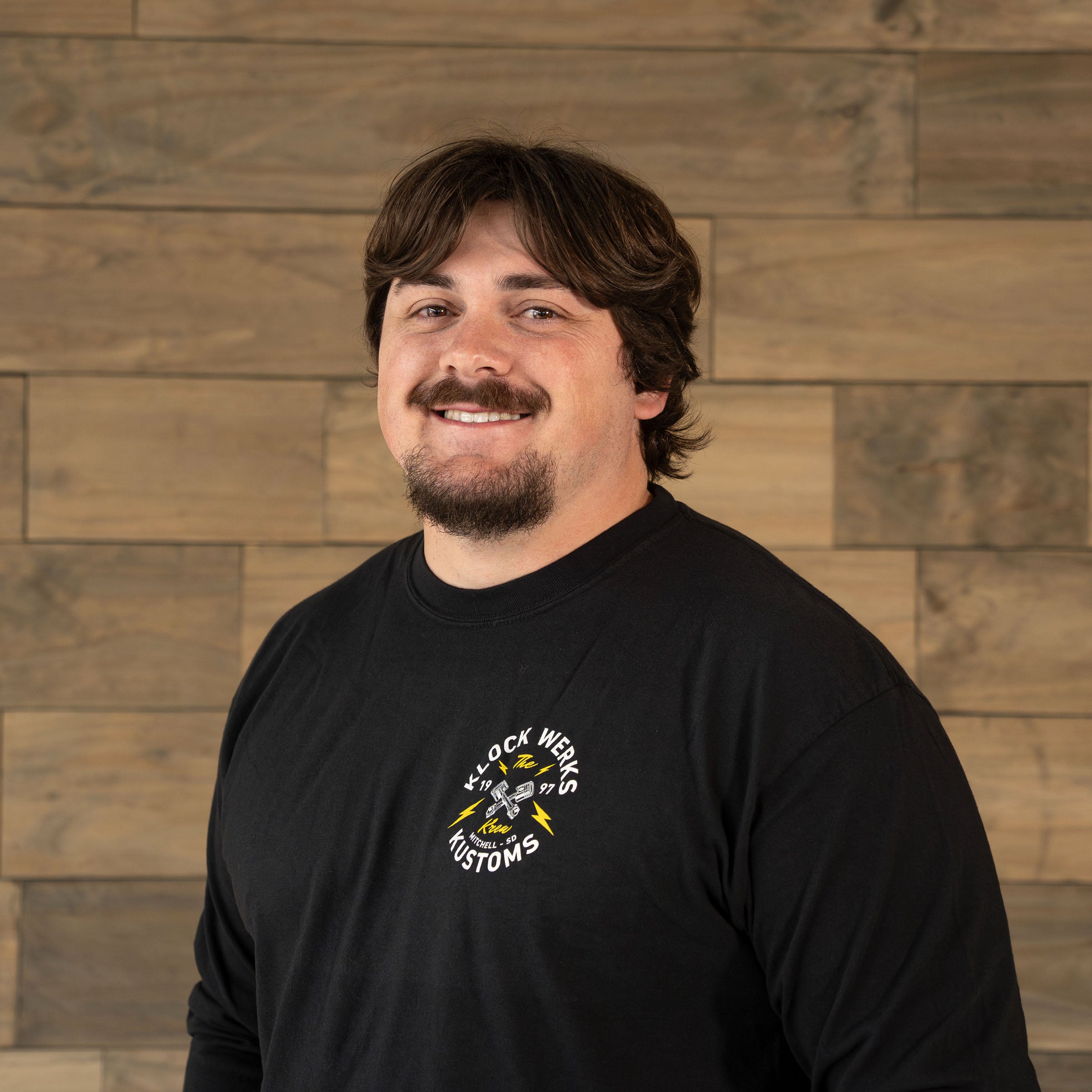 Trevor Wilkinson of Klock Werks poses in front of a wooden backdrop for his employee photo.