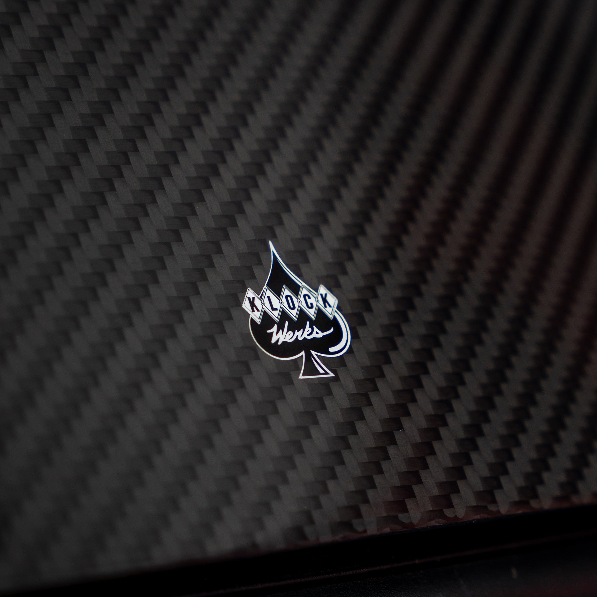 A close-up of a glossy black carbon fiber textured surface with a small silver and white spade-shaped logo in the center reading 'Klock Werks'.