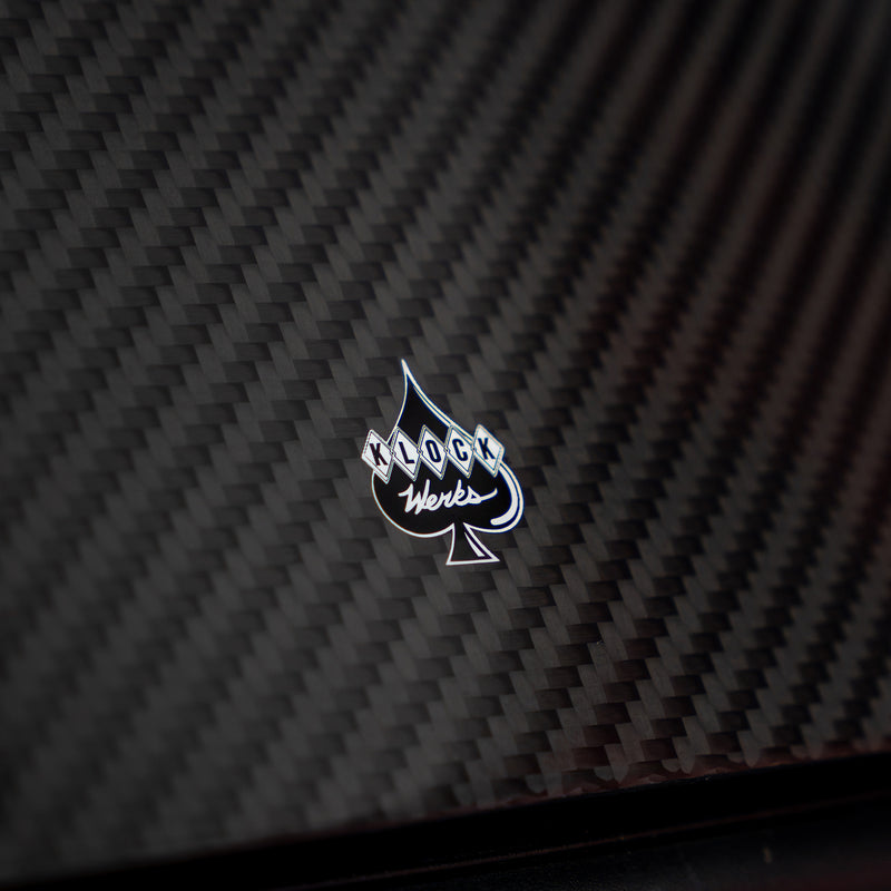 A close-up of a glossy black carbon fiber textured surface with a small silver and white spade-shaped logo in the center reading 'Klock Werks'.