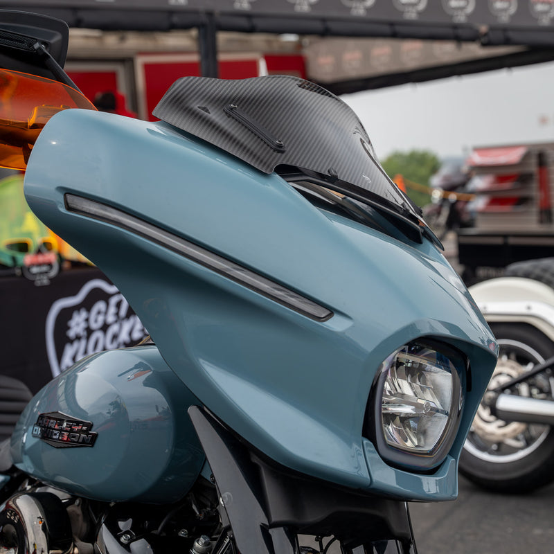 Close-up of a light blue 2023.5+ Harley-Davidson Street Glide front fairing with a black carbon-fiber-look windscreen and a single round headlight, photographed at an outdoor event.(8" Carbon Fiber)