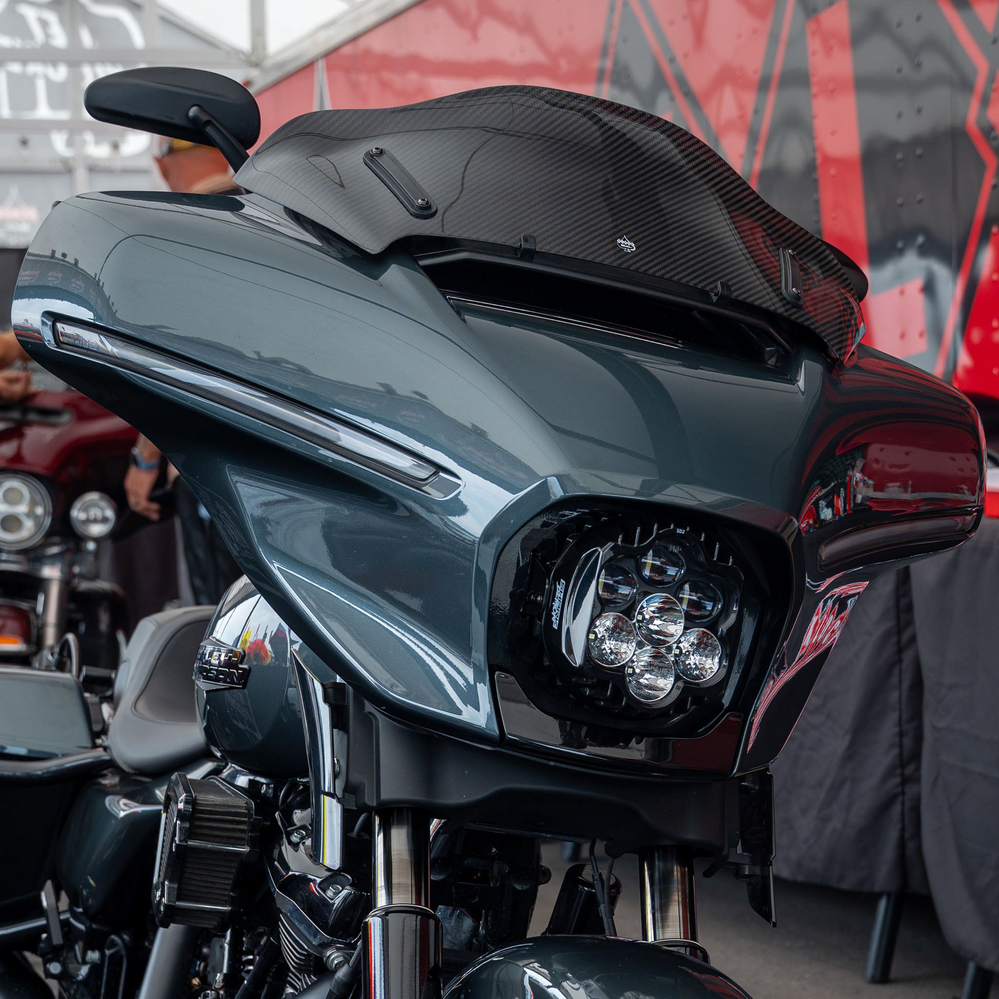 Close-up of a dark metallic gray Harley-Davidson Street Glide touring motorcycle with a black carbon-fiber Flare® Windshield and LED headlight, photographed at an outdoor event with a red and black banner in the background. (8" Carbon Fiber)
