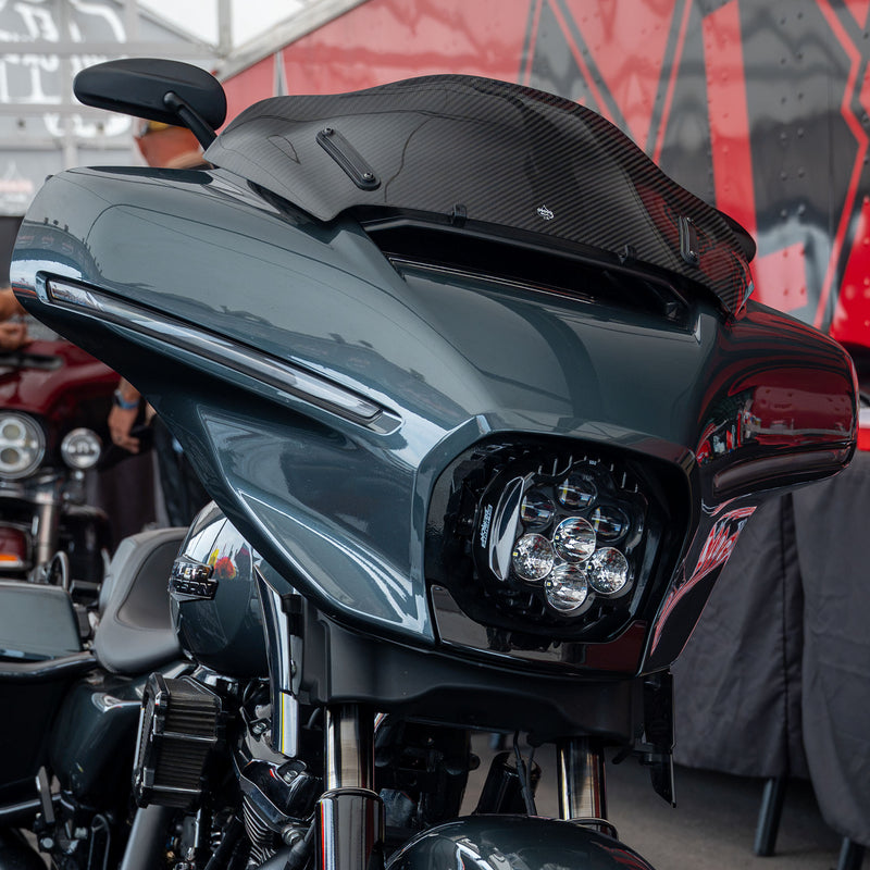Close-up of a dark metallic gray Harley-Davidson Street Glide touring motorcycle with a black carbon-fiber Flare® Windshield and LED headlight, photographed at an outdoor event with a red and black banner in the background. (8" Carbon Fiber)