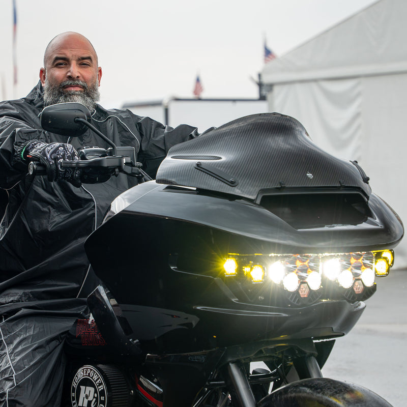 A man wearing a black raincoat and gloves sits on a black custom Harley-Davidson® Road Glide motorcycle with a 10" carbon fiber Flare® Windscreen and bright LED lights, photographed outdoors in the rain. (10" Carbon Fiber)