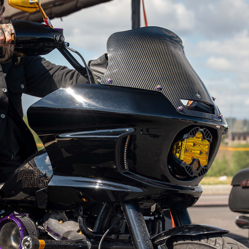 Close-up of a black Harley-Davidson motorcycle with a carbon-fiber-textured windscreen and a bright yellow headlight, shown outdoors with a rider's hand visible on the handlebar. (8" Carbon Fiber)
