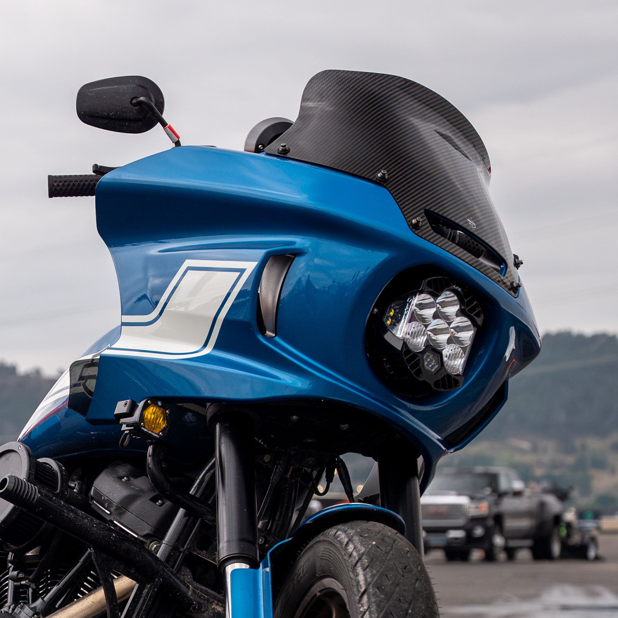 Close-up of a blue motorcycle with a large black carbon-fiber-style windscreen and a cluster of LED headlights, photographed outdoors with a blurred overcast sky and distant vehicles in the background. (8" Carbon Fiber)