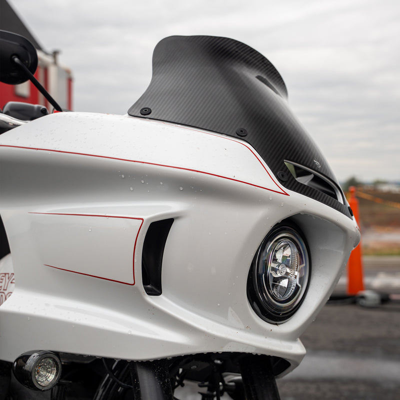 Close-up of a white motorcycle front fender and windscreen with red pinstripes, a black carbon-fiber-look windscreen, and a round headlight, photographed outdoors on a wet paved surface. (8" Carbon Fiber)