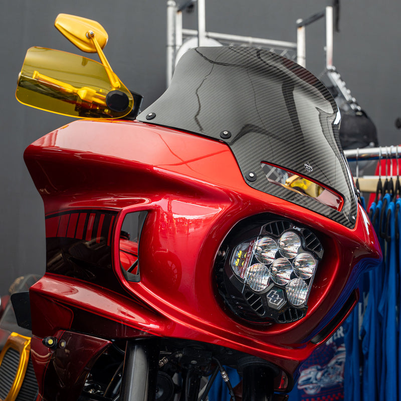 Close-up of a red motorcycle front fairing with a large round LED headlight, a black carbon-fiber-look windshield, and a yellow-tinted side mirror, photographed in a workshop setting. (8" Carbon Fiber)