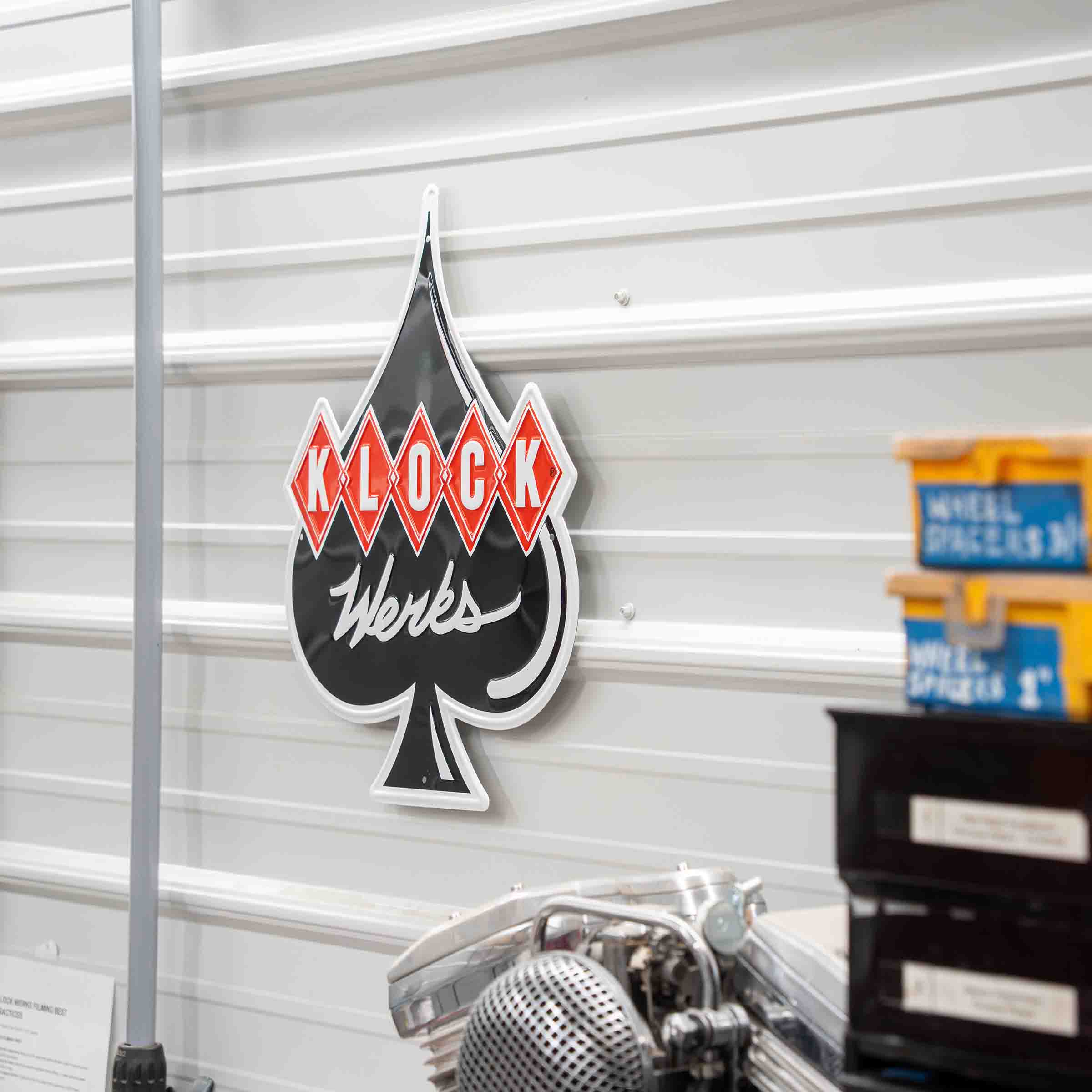 A Klock Works branded black and white spade-shaped wall sign with red diamond accents, mounted on a white corrugated metal wall in a workshop setting.