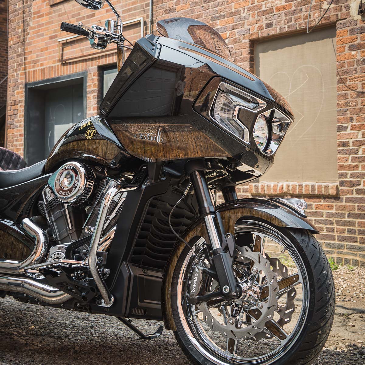 A close-up shot of a custom black and dark wood grain motorcycle parked outdoors in front of a brick building.